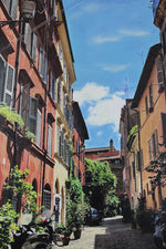 quintessential European street scene. Pale pink yellow buildings, cobblestones and scooters and lush green trees make up up this wonderful scene with blue sky.